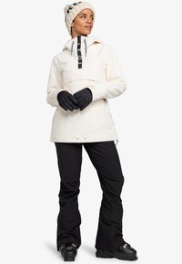 Leichte Cremepulloverjacke mit einer Fronttasche, grauen Akzenten und einem schwarzen Logo; kombiniert mit schwarzen isolierten Skihosen und Handschuhen.