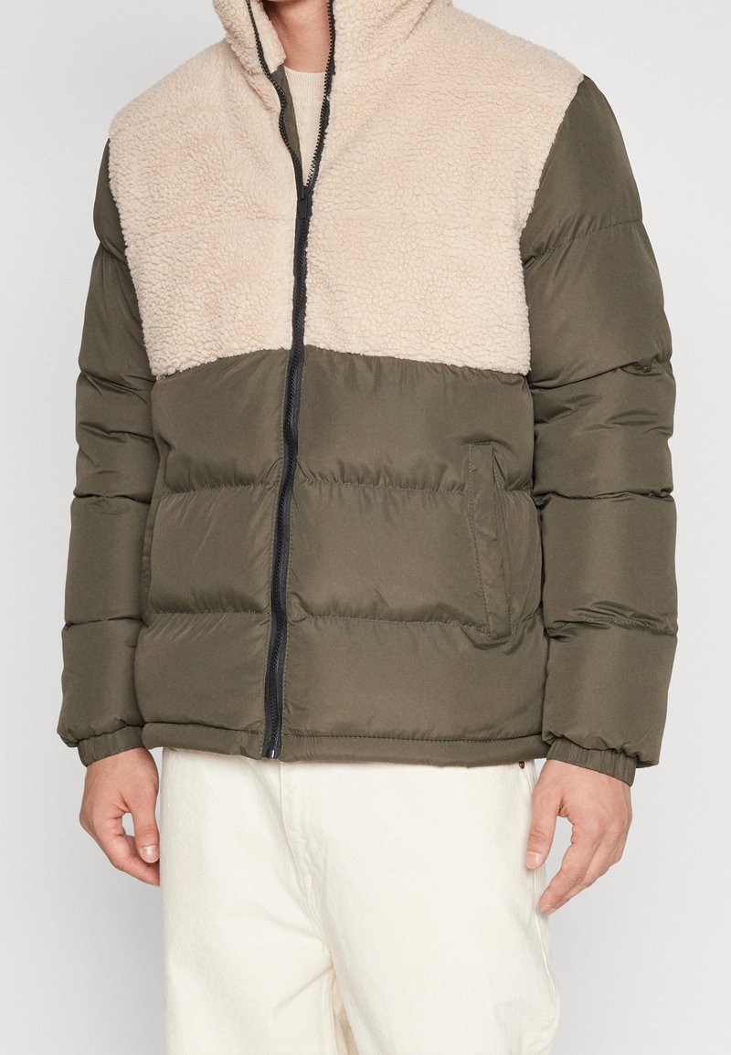 Homme portant une veste zippée en polaire olive et beige avec des poignets élastiques et un pantalon blanc, montré du cou aux hanches.