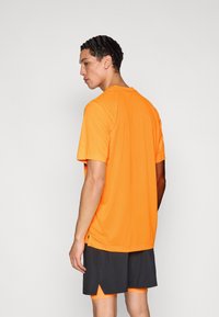 Πορτοκαλί αθλητική μπλούζα με κοντά μανίκια, λεία υφή, στρογγυλή λαιμόκοψη και πλαϊνές χαρακιές, συνδυασμένη με σκούρα σορτς.