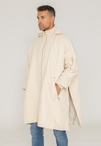Beżowy, oversize płaszcz z wysokim kołnierzem, zamkiem błyskawicznym, bocznymi kieszeniami i fakturowanym rękawem. Wykonany z gładkiego, lekkiego materiału.