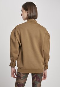 Hanorac maro cu guler înalt, mâneci largi și mansete elastice, purtat cu colanți de camuflaj care au nuanțe de verde și burgund.