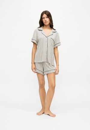 Jeune femme debout pieds nus portant un pyjama blanc à motifs avec manches courtes boutonné et un short assorti sur un fond uni.