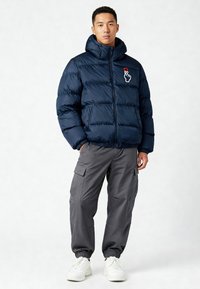 Giacca imbottita blu navy con collo alto, chiusura con cerniera e tasche frontali. Presenta una grafica di una mano e un cuore sul petto. Indossata con pantaloni cargo grigi.