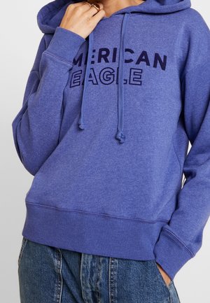 Paarse hoodie met "AMERICAN EAGLE" in donkerblauw gedrukt. Beschikt over een trekkoordkap en ribgebreide manchetten en zoom. Zachte materiaalkwaliteit.