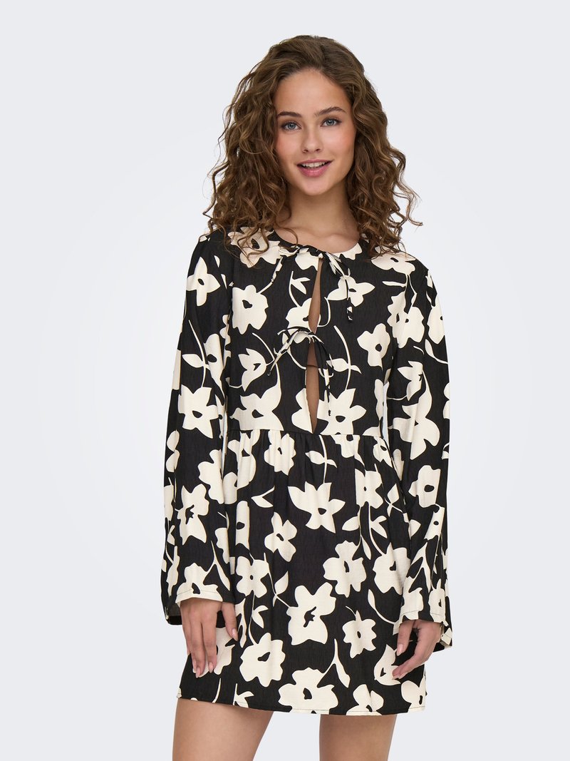 Jeune femme aux cheveux bouclés portant une robe noire à manches longues avec de grands motifs floraux blancs et des découpes en forme de goutte nouées au niveau de l’encolure et de la poitrine.