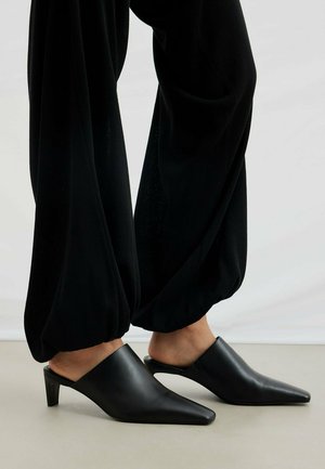 Persoon draagt zwarte puntige muiltjes met hak en losse zwarte broek met elastische boorden, staande op een lichte vloer.