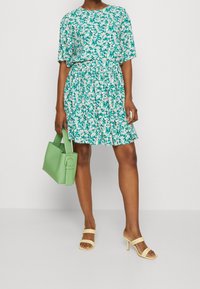 Vestido floral en turquesa y blanco, con mangas cortas, cintura fruncida y falda acampanada. Combinado con un bolso verde y sandalias de tiras amarillas claras.