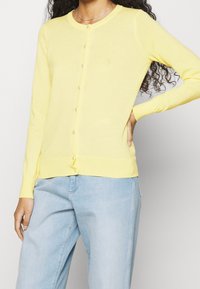 Gul stickad cardigan med rund hals, knäppning fram och långa ärmar. Detaljerad med små knappar och en åtsittande nederkant.