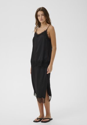 Mlada ženska stoji, oblečena v črno brezrokavnik z čipko obrobo in ujemajočo midi krilo s stranskim razporkom, črne natikače, nevtralna podlaga.