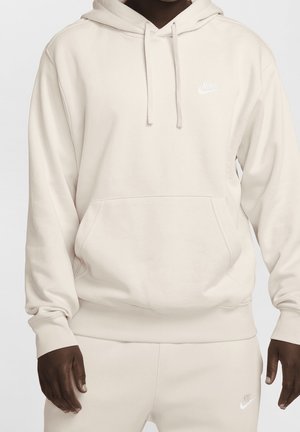 Person iført cremefarvet Nike-hættetrøje og matchende joggingbukser med hvide Nike-logoer på bryst og lår.