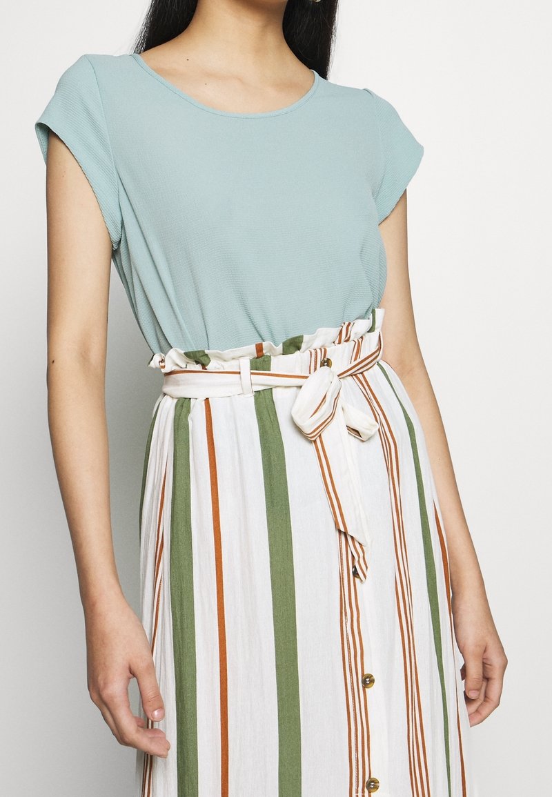 Femme portant un haut bleu clair à manches courtes rentré dans une jupe blanche taille haute avec des rayures verticales vertes et orange et une ceinture nouée.