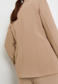 Blazer beige sur mesure avec épaules structurées, tissu lisse et coutures marquées, vu de dos. Design simple, sans éléments métalliques visibles.
