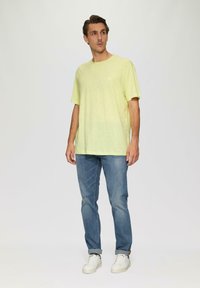 Hellgelbes T-Shirt mit kurzen Ärmeln, Rundhalsausschnitt und subtiler Textur, kombiniert mit blauen Jeans und weißen Sneakers. Lässiger Schnitt.