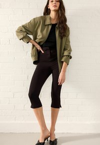 Olivegrüner oversized Blazer mit Kragen, schwarzes Top darunter und figurbetonte, dunkelbraune Capris. Schwarze, hochhackige Mules runden das Outfit ab.