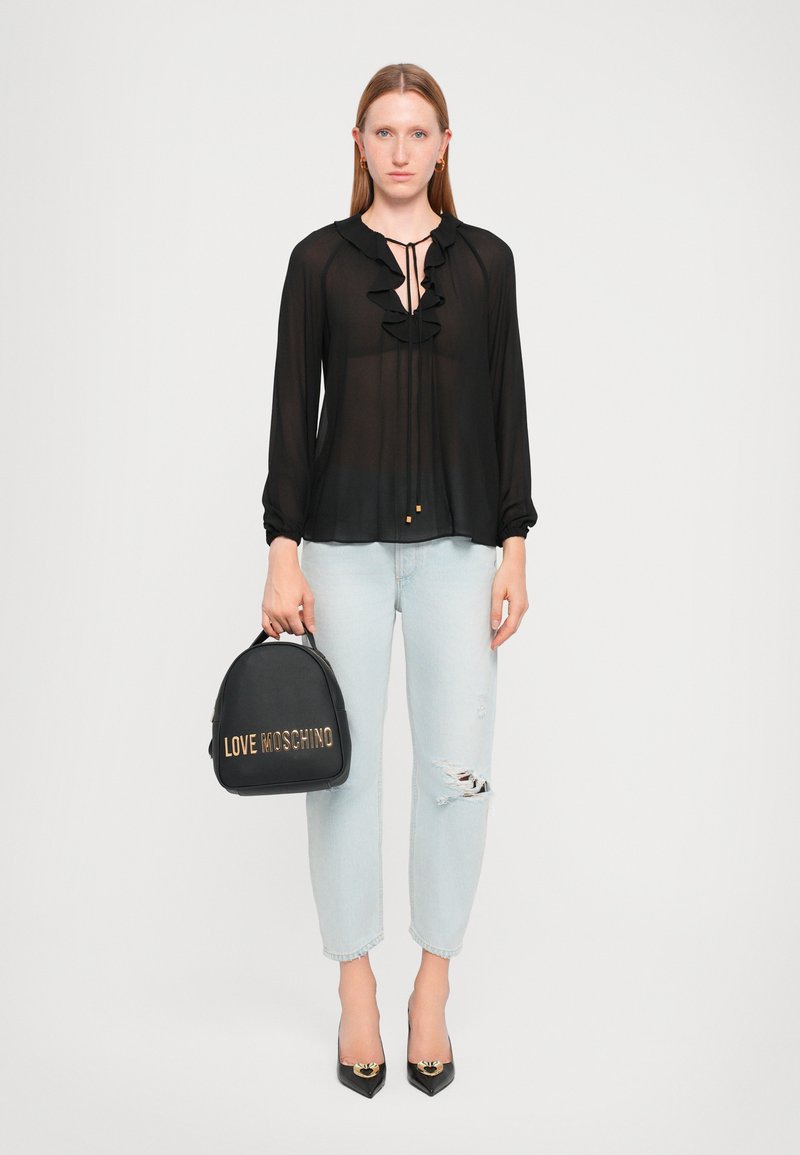 Schwarze transparente Bluse mit Rüschen, hellblaue verwaschene Jeans und eine schwarze Handtasche mit goldenen "LOVE MOSCHINO"-Logo. Schwarze spitze Absatzschuhe.