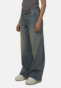 Brede blauwe spijkerbroek met vervaagde details. Beschikt over vijf zakken, riemlussen en een klassieke knoop- en ritsluiting. G gedragen met witte sneakers.