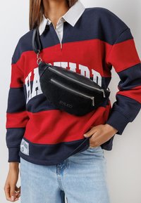 Črn fanny pack iz blaga z dvema zadrgama, pritrjenim trakom in embosiranim logotipom, nošen preko rdeče in mornarsko modre pruge.