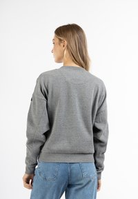 Felpa grigia con colletto a girocollo, orlo e polsini a coste. Presenta spalle cadenti e una texture liscia. Indossata sopra jeans blu.