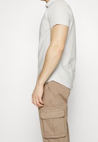 Homme portant une chemise à manches courtes boutonnée gris clair et un pantalon cargo beige, debout de profil devant un fond blanc uni.