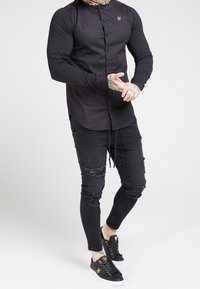 Hombre vistiendo una camisa negra ajustada con botones y un logo dorado, jeans ajustados negros desgastados y zapatillas negras con acentos dorados, de pie.