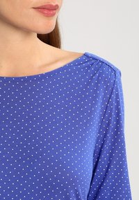 Blå långärmad tröja i mjukt material, med rundad halsringning och små vita triangelmönster över hela, med åtstramade axlar.