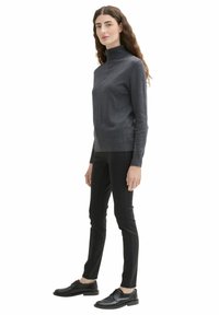 Grauer Rollkragenpullover aus weichem Stoff, kombiniert mit schwarzen Slim-Fit-Hosen und schwarzen Schnürschuhen. Das Modell steht im Profil.