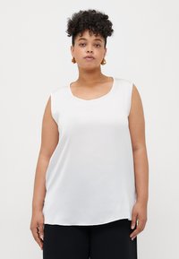 Top bianco senza maniche realizzato in un tessuto liscio; presenta un collo rotondo e un orlo leggermente curvo. Indossato con pantaloni neri.