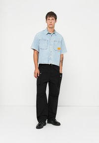 Ung mand iført en lyseblå cropped denimskjorte med lommepatch og sorte brede cargo bukser, der står foran en hvid baggrund.
