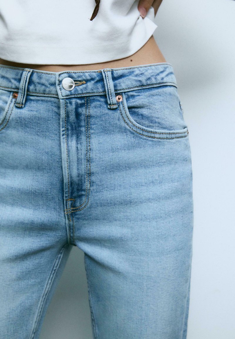 Ljust blå högmidjade jeans i denim med en klassisk femfickdesign, mässingsknapp och subtila blekningar på tyget.