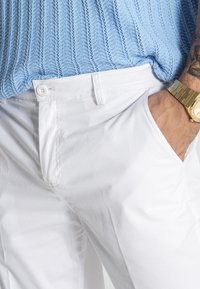 Weiße Baumwollhose mit geradem Schnitt, ausgestattet mit zwei Seitentaschen, einem Knopfverschluss und einer flachen Vorderseite. Getragen mit einem hellblauen Strickoberteil.