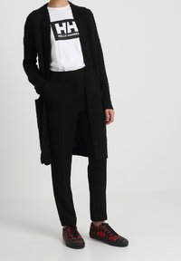 Musta, teksturoitu pitkä kardiganin päälle valkoinen t-paita, jossa on logo. Mustat räätälöidyt housut ja punaiset ruutukuosiset lenkkarit täydentävät asua.