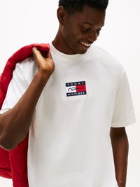 Balta medvilnės marškinėliai su stačiakampiu daugiaspalviu logotipo pleistru, ant kurio užrašyta "TOMMY HILFIGER." Subjektas laiko raudoną siuvinėtą striukę per vieną petį.