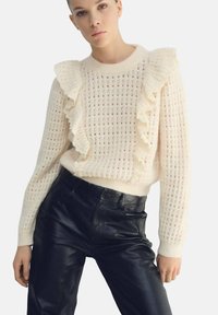 Suéter de punto color crema con cuello con volantes y patrón texturizado, combinado con pantalones de cuero negros de talle alto.