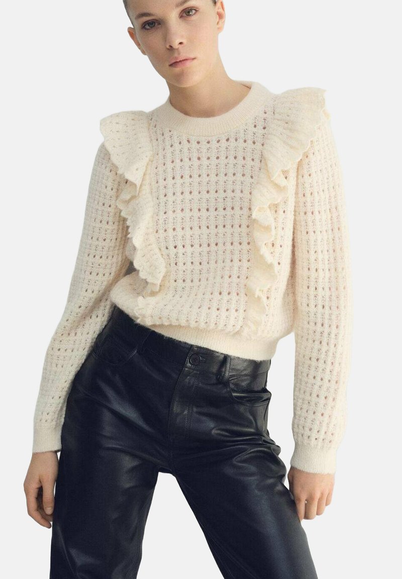 Suéter de punto color crema con cuello con volantes y patrón texturizado, combinado con pantalones de cuero negros de talle alto.