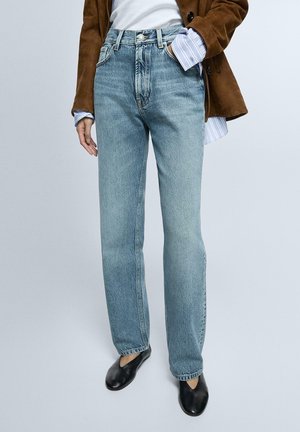 Persona che indossa jeans azzurri a vita alta con gamba dritta, top bianco, giacca marrone e scarpe slip-on nere, in piedi davanti a uno sfondo semplice.