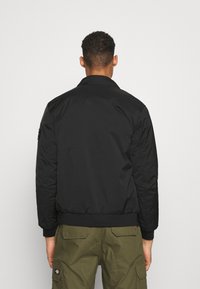 Veste bomber noire avec un col et des poignets côtelés, présentant une texture douce et mate et un ourlet droit, portée avec un pantalon cargo vert olive.