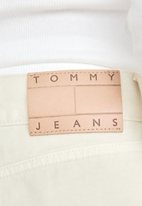 Lys beige denim med et brunt lærmerke preget med "TOMMY JEANS." Merket er rektangulært med en sydd kant.