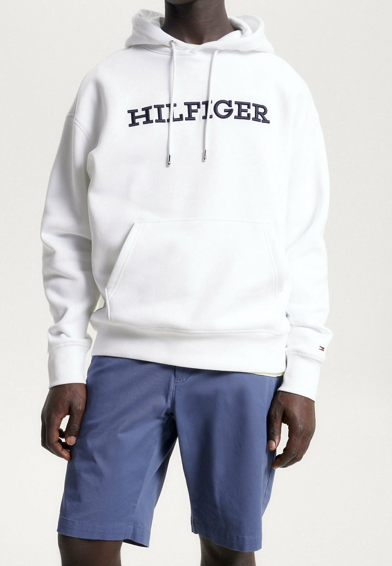 Homme portant un sweat à capuche blanc avec le texte "HILFIGER" et un short bleu jusqu'aux genoux, debout devant un fond clair uni.