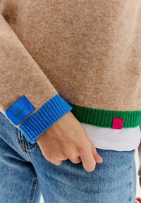 Sweter wykonany z włóczki w jasnobrązowym kolorze, z niebieskimi i zielonymi ściągaczami. Posiada teksturowany materiał oraz małe naszywki z logo w kolorze niebieskim i różowym.