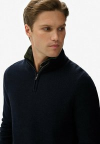Marineblå zip-up sweater med et tekstureret mønster, der har en ståkrave lagdelt over en grønternet skjorte nedenunder.