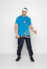 Blå kortermet t-skjorte med en grafisk sirkeldesign, sammen med marineblå joggebukser. Han holder en hvit tennisracket og har på seg svarte joggesko.