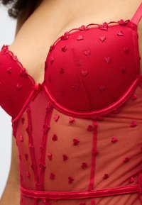 Κόκκινο εσώρουχο με καρδιόσχημες κεντητές λεπτομέρειες σε διάφανο ύφασμα, με αντίθετες μονόχρωμες κόκκινες λεπτομέρειες και λεπτά κυματιστά τελειώματα.
