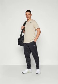Bež Nike t-särk, mustad Nike spordipüksid, hallid Nike jooksujalanõud, mustat spordikotiga, seistes valgel taustal.