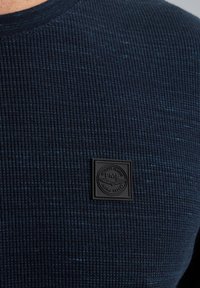 Marineblauer, gerippter Pullover aus strukturiertem Stoff mit dezenten blauen Sprenkeln. Mit einem auffälligen schwarzen Logopatch auf der Brust.