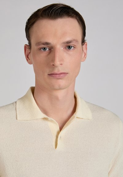 Młody mężczyzna z krótkimi brązowymi włosami, ubrany w kremową, teksturowaną koszulę z kołnierzykiem, zwrócony przodem, z neutralnym wyrazem twarzy na jednolitym szarym tle.