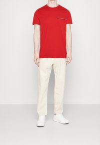 T-shirt in cotone rosso con maniche corte e collo rotondo, caratterizzato da una grafica testuale nera nella zona della tasca sul petto. Pantaloni beige a gamba dritta.