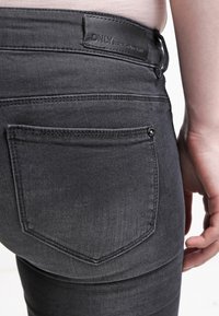 Grå denimjeans med en slät yta, med en läderetikett och en bakficka med dekorativ sömnad och knappdetalj.