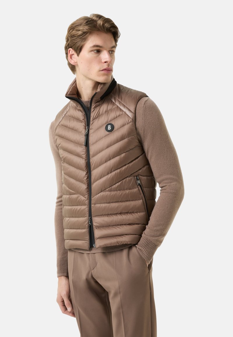 Braune Steppweste mit Reißverschluss vorne und Chevron-Muster, ausgestattet mit einem schwarzen Logoabzeichen und Seitentaschen, getragen über einem hellbraunen Pullover.