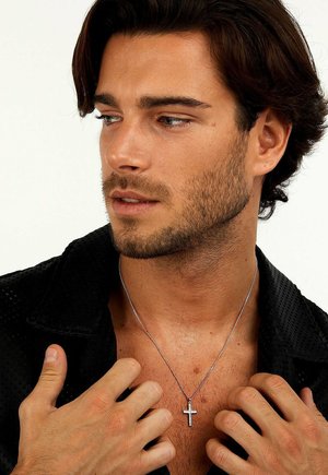 Mann mit kurzem Bart und dunklem Haar, der ein offenes schwarzes Hemd und eine silberne Kreuzkette trägt, hält mit beiden Händen den Hemdkragen fest.