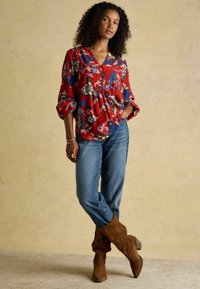 Rote Bluse mit floralem Muster, V-Ausschnitt und lockeren Ärmeln, kombiniert mit blauen Jeans und braunen Wildleder-Stiefeln bis zum Knie. Strukturierter Hintergrund.